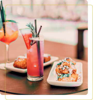 Un tavolo apparecchiato con drink colorati, tra cui un cocktail rosso e uno arancione, accompagnati da deliziosi antipasti fritti. Un'immagine che trasmette un'atmosfera vivace e festiva.