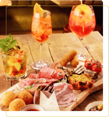 Tavola imbandita con diversi antipasti, salumi, formaggi e cocktail colorati, su un tavolo in legno. Atmosfera accogliente.