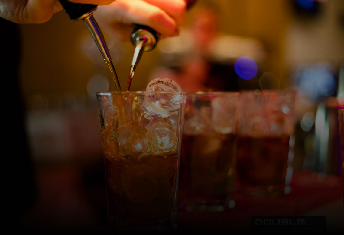Barman versa attentamente una bevanda in un bicchiere. Dettagli sul ghiaccio e altri drink sullo sfondo. Atmosfera vivace e festosa del bar.