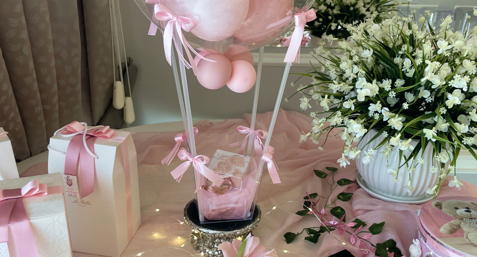Decorazione per un evento festivo con palloncini rosa, nastri e un bouquet di fiori bianchi, su un tavolo elegante.