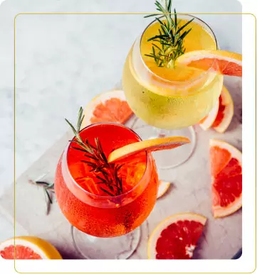 Aperitivo. Due bicchieri di cocktail colorati, uno rosso e uno giallo, decorati con fette di agrumi e rosmarino, su un tavolo di marmo.