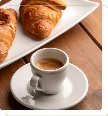 Un caffè espresso fumante in una tazzina bianca, posizionato accanto a un piatto con croissant dorati su un tavolo di legno.