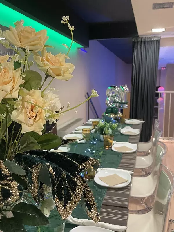 Vista di un tavolo elegantemente decorato per una festa, con fiori gialli, stoviglie bianche e palloncini argentati. L'atmosfera è festiva e colorata.