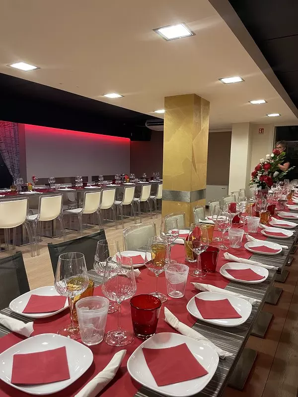 Interno di un ristorante elegante, tavoli imbanditi con tovaglie rosse e bicchieri di vetro. Mentre sullo sfondo si vedono sedie bianche e un'illuminazione calda, un membro del personale sistema un bouquet di rose.