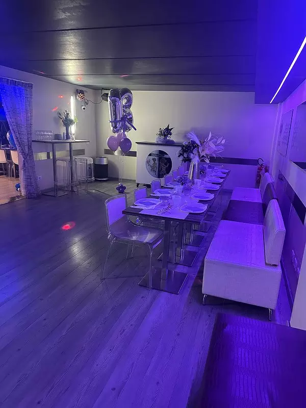 Interno di un locale con tavolo elegante servito per un evento, illuminazione romantica e decorazioni floreali in un'atmosfera festiva.