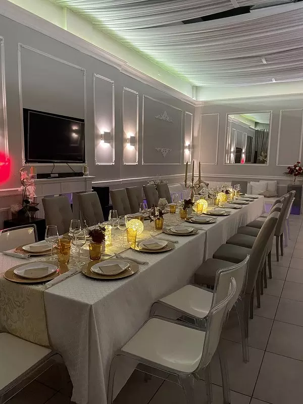Immagine di una elegante tavola apparecchiata per una cena, con tovaglia bianca, piatti dorati, bicchieri e candelabri luminosi, in un ambiente raffinato e ben illuminato.