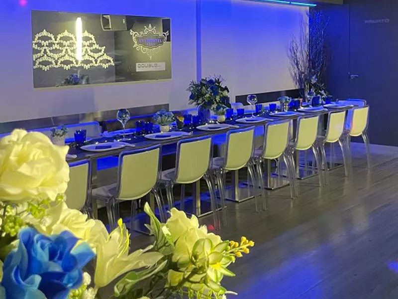 Ristorante elegante con tavolo lungo e apparecchiato, illuminato da luci blu. Centrotavola floreali e decorazioni raffinate.