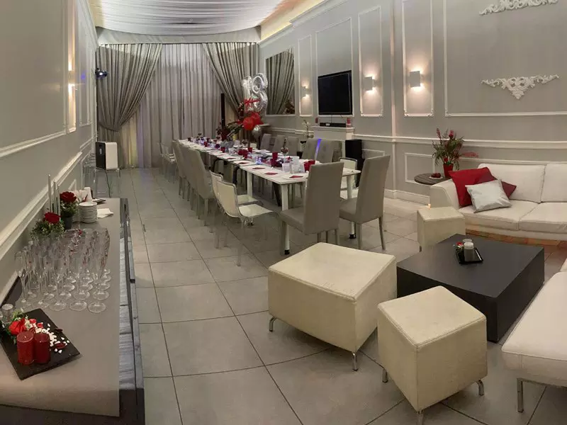 Interno elegante di un locale con tavolo lungo allestito per una cena. Mobili moderni, decorazioni floreali e dettagli raffinati.