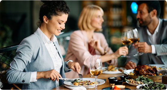 Una donna in giacca grigia gusta un piatto delizioso in un ristorante elegante, mentre due amici brindano. Atmosfera conviviale.